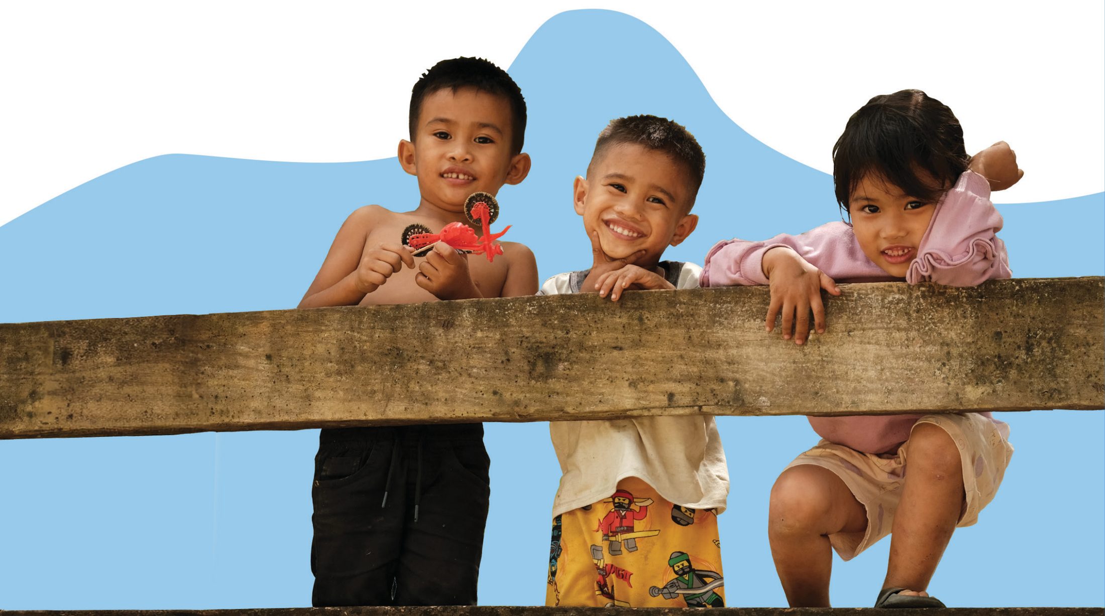Three children smile over a wooden fence